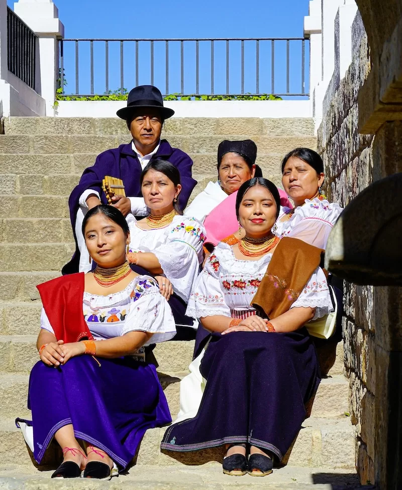 A multi-generational family in traditional highland dress poses on stone steps in Otavalo. Across Ecuador, family extends beyond the household. Cousins, godparents, and neighbors form a web of everyday support that keeps community life vibrant.