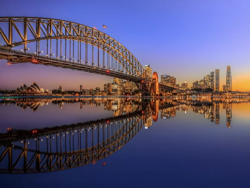 Sydney Harbour Bridge at sunset with Opera House and city skyline reflected in water showing Australia's iconic architecture and stable environment for expats