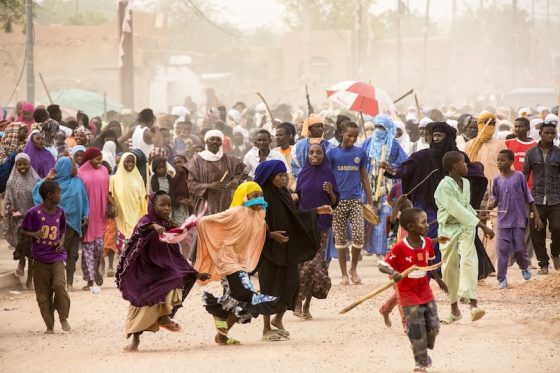 Large crowd of African families and children in colorful clothing gathered in dusty settlement, representing humanitarian crisis and displaced communities from regional conflicts and security crises