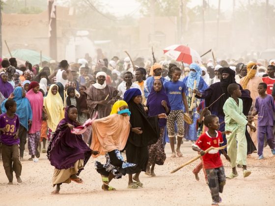 Large crowd of African families and children in colorful clothing gathered in dusty settlement, representing humanitarian crisis and displaced communities from regional conflicts and security crises