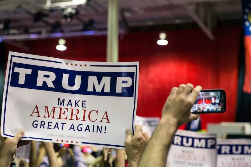 Political rally crowd holding Trump campaign sign, representing US political messaging and foreign policy rhetoric discussed in global news coverage