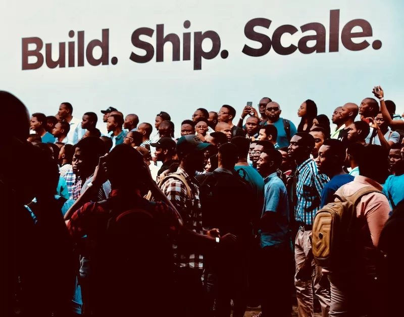 Diverse crowd of African people gathered at community event with "Build. Ship. Scale." banner, representing economic development, fintech innovation, and digital transformation in African communities