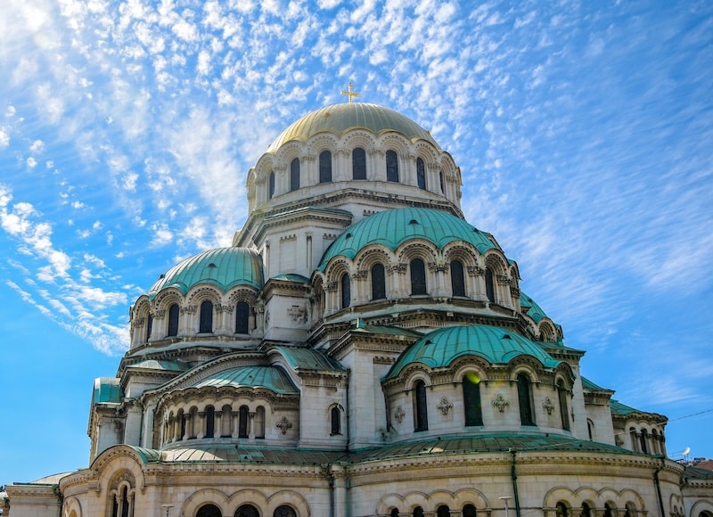History rises above you in Sofia. Copper domes catch the sun. A cathedral roof curves like waves frozen in stone. You stand on a street where centuries have stacked themselves into the skyline. Bulgaria does not hide its past behind velvet ropes. It asks you to walk right through it.