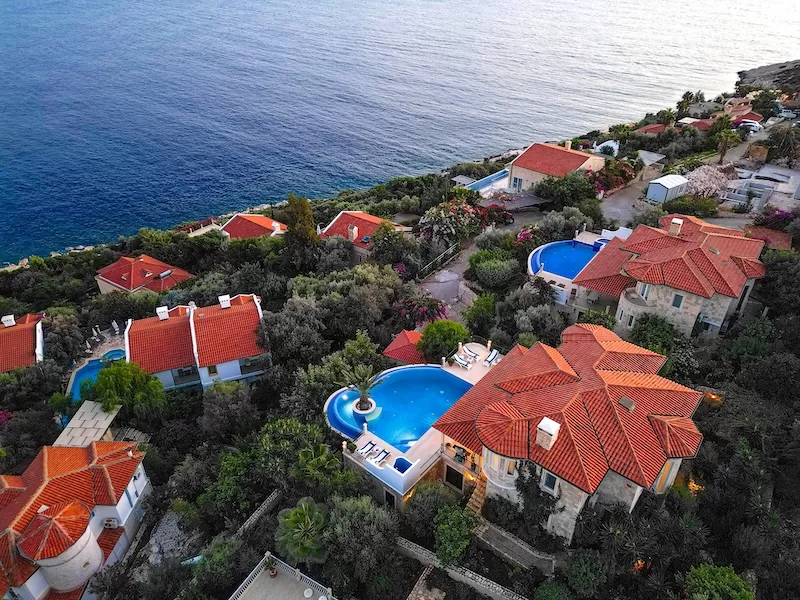 Aerial view of luxury Mediterranean coastal properties with red tile roofs and blue swimming pools overlooking turquoise sea, representing real estate investment and wealth building through global property
