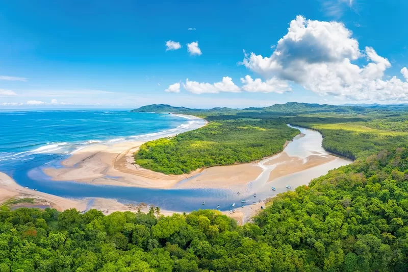 An aerial view of a winding river meeting the turquoise Pacific Ocean at a lush, tropical beach in Costa Rica, showing the natural rhythm of coastal life.