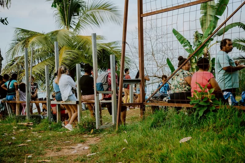 A group of locals and travelers gather behind a rustic fence on a grassy hill, overlooking a tropical landscape and sharing a moment of community connection.