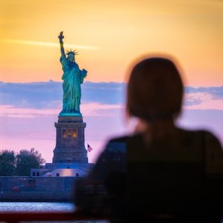 Woman viewing Statue of Liberty at sunset overlooking New York Harbor symbolizing U.S. immigration and global citizenship concerns during nationwide protests