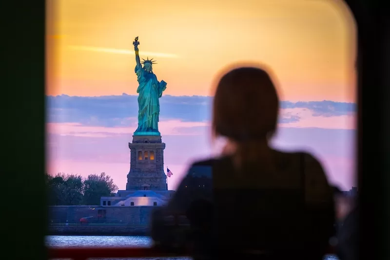 Woman viewing Statue of Liberty at sunset overlooking New York Harbor symbolizing U.S. immigration and global citizenship concerns during nationwide protests