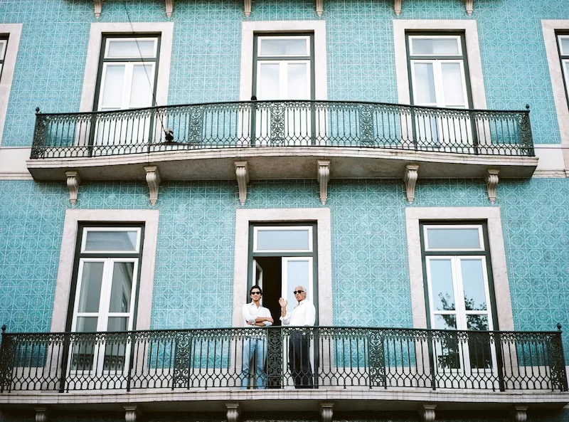 Two men stand on a balcony of a building with a striking blue tiled facade, representing the expat experience in popular European cities like Lisbon where housing costs are a primary driver of inflation