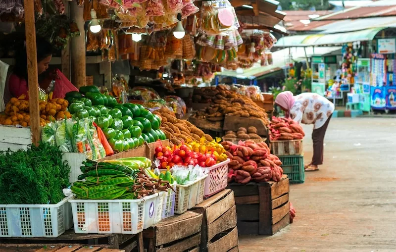 A bustling outdoor market in Southeast Asia, with a woman in a hijab browsing fresh vegetables, showcasing the vibrant local life that exists beyond tourist areas.