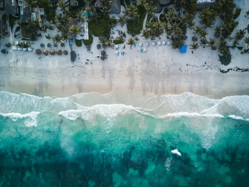 High-angle drone shot of a pristine turquoise ocean meeting a white sand beach lined with palm trees, illustrating the allure of tourist hotspots like Tulum that are now experiencing significant "Tourist Inflation."
