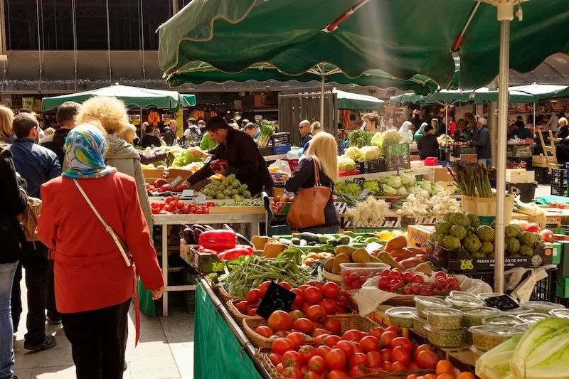A vibrant, busy local market filled with people and stalls overflowing with fresh, locally-grown vegetables and produce, highlighting the "Adaptation Strategy" to hedge against imported food inflation.