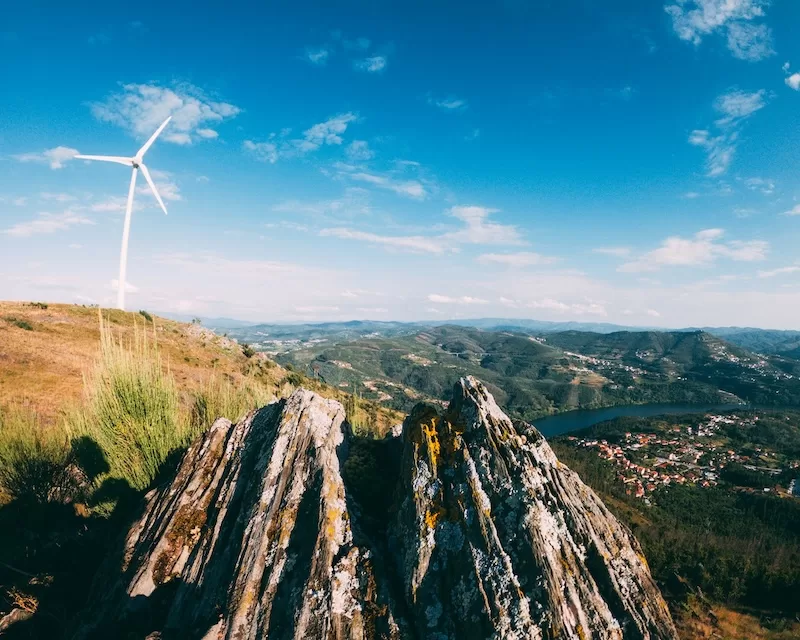 A wind turbine on a hill overlooking a river and village in Portugal, symbolizing the country's commitment to renewable energy and sustainable development.