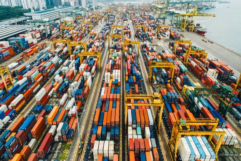 An expansive aerial view of a massive, colorful shipping container port, representing the global supply chain and the "Habit Tax" of imported goods that are subject to high container shipping costs and trade friction.