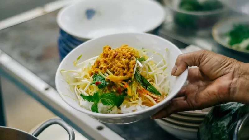 A hand holds a bowl of Num Banh Chok, a traditional Khmer noodle dish, representing the delicious and affordable local food in Cambodia for budget travelers