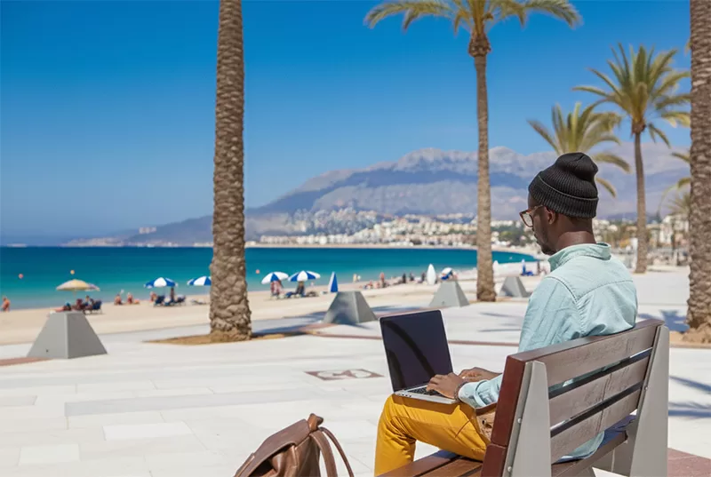 A man wearing a beanie works on his laptop on a bench overlooking a sunny beach, a perfect example of a bleisure lifestyle that blends work and relaxation.