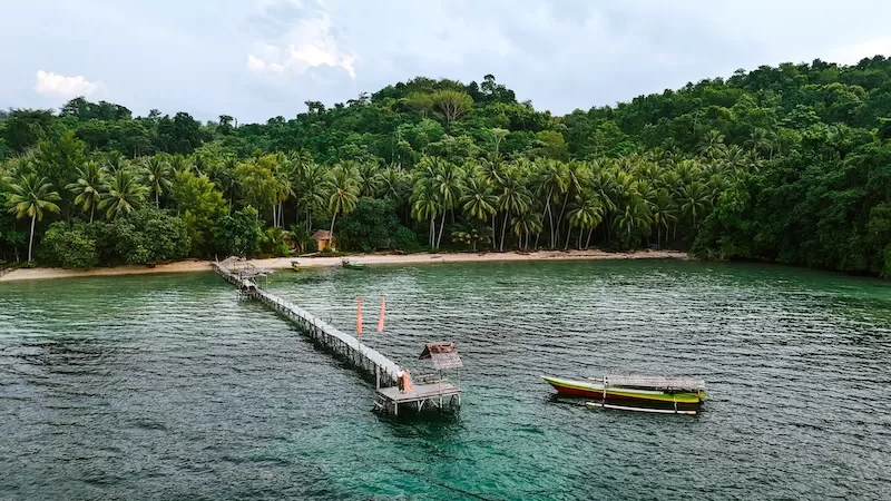 A wooden dock extends into turquoise water with a traditional fishing boat, representing the pristine natural beauty and slow travel opportunities in tropical destinations