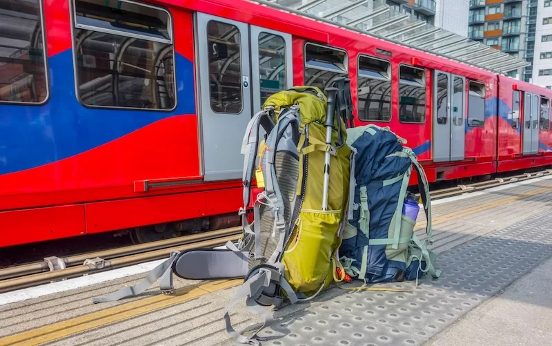 Two large backpacks on a train platform next to a red train, symbolizing the use of public transportation for long-term, sustainable travel on a budget.