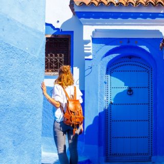 A woman with a leather backpack walks through a vibrant, bright blue-painted alleyway, capturing the iconic aesthetic of cities like Chefchaouen.