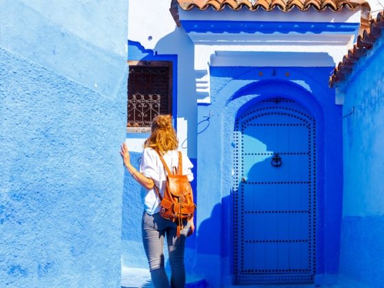 A woman with a leather backpack walks through a vibrant, bright blue-painted alleyway, capturing the iconic aesthetic of cities like Chefchaouen.