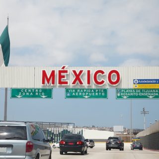Mexican flag flying above a large red MEXICO sign at a border crossing with vehicles and road infrastructure visible under a bright blue sky above