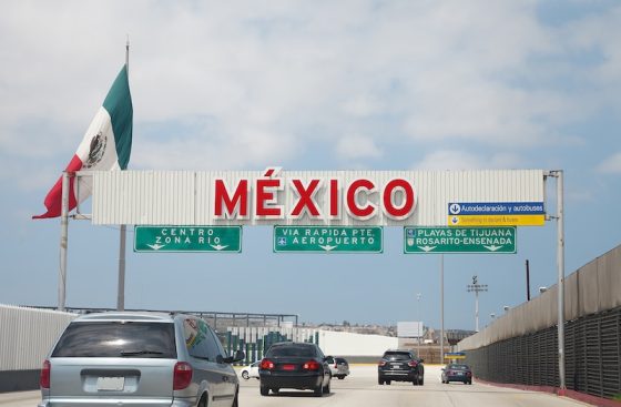 Mexican flag flying above a large red MEXICO sign at a border crossing with vehicles and road infrastructure visible under a bright blue sky above