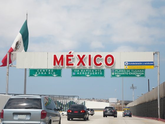 Mexican flag flying above a large red MEXICO sign at a border crossing with vehicles and road infrastructure visible under a bright blue sky above