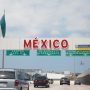Mexican flag flying above a large red MEXICO sign at a border crossing with vehicles and road infrastructure visible under a bright blue sky above