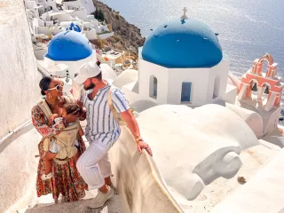 A young family stands on a white stone staircase in Santorini, Greece, with iconic blue-domed churches and the sparkling Aegean Sea in the background.