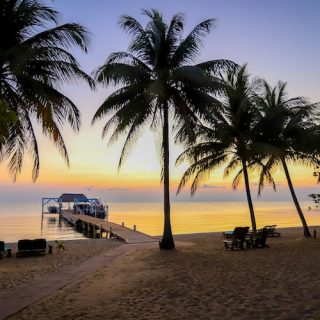 A long wooden pier extends into the calm Caribbean Sea at sunset in Belize, with silhouettes of palm trees and lounge chairs on the sandy beach shore.