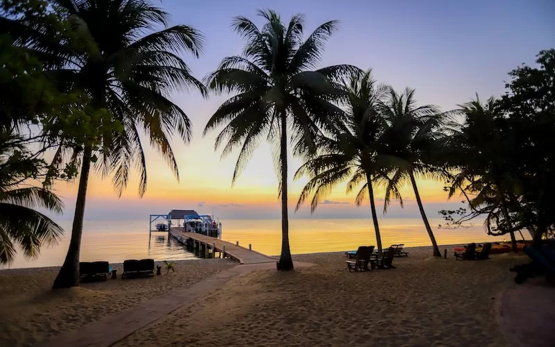 A long wooden pier extends into the calm Caribbean Sea at sunset in Belize, with silhouettes of palm trees and lounge chairs on the sandy beach shore.