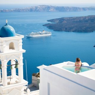 A woman relaxes in a private pool on a white-washed cliffside in Santorini, Greece, overlooking the deep blue Aegean Sea with a cruise ship and distant islands under a clear sky.