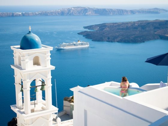A woman relaxes in a private pool on a white-washed cliffside in Santorini, Greece, overlooking the deep blue Aegean Sea with a cruise ship and distant islands under a clear sky.