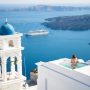 A woman relaxes in a private pool on a white-washed cliffside in Santorini, Greece, overlooking the deep blue Aegean Sea with a cruise ship and distant islands under a clear sky.