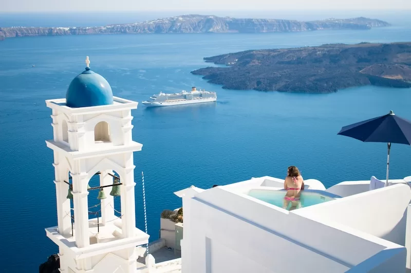 A woman relaxes in a private pool on a white-washed cliffside in Santorini, Greece, overlooking the deep blue Aegean Sea with a cruise ship and distant islands under a clear sky.