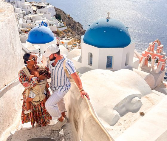 A young family stands on a white stone staircase in Santorini, Greece, with iconic blue-domed churches and the sparkling Aegean Sea in the background.