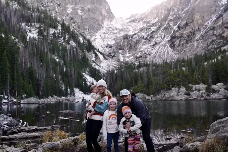 A family of five, including a baby in a carrier, stands together on a rocky shore beside a serene lake, with a dramatic backdrop of snow-dusted mountains and evergreen trees.