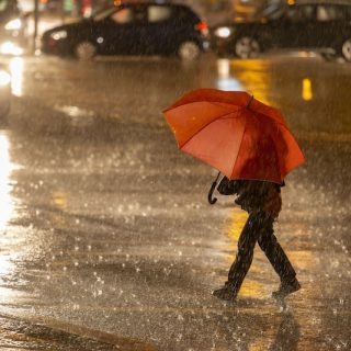 A lone person holding a bright red umbrella walks across a wet city street at night, with car headlights and reflections creating a moody, urban scene.