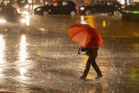 A lone person holding a bright red umbrella walks across a wet city street at night, with car headlights and reflections creating a moody, urban scene.