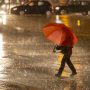A lone person holding a bright red umbrella walks across a wet city street at night, with car headlights and reflections creating a moody, urban scene.