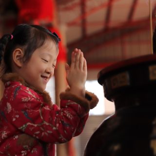 A young East Asian girl with braided hair and a red traditional jacket smiles with her eyes closed, pressing her hands together to pray at a temple.