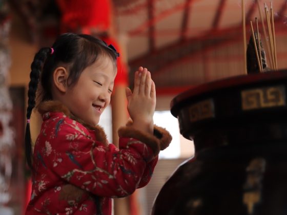 A young East Asian girl with braided hair and a red traditional jacket smiles with her eyes closed, pressing her hands together to pray at a temple.