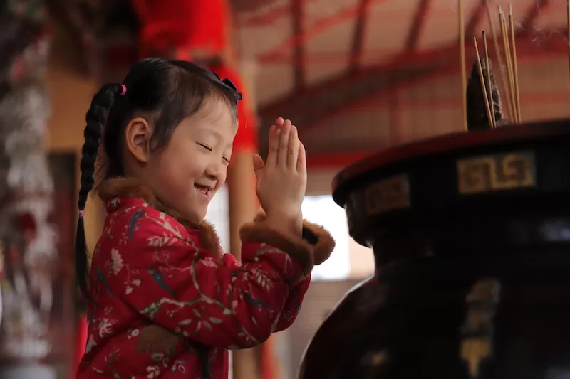 A young East Asian girl with braided hair and a red traditional jacket smiles with her eyes closed, pressing her hands together to pray at a temple.