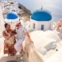 A young family stands on a white stone staircase in Santorini, Greece, with iconic blue-domed churches and the sparkling Aegean Sea in the background.