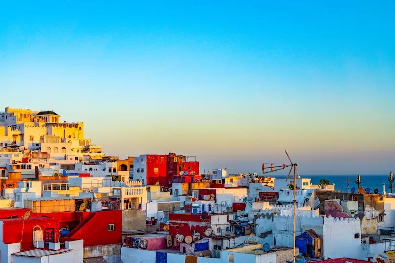 A panoramic view of a Moroccan hillside city with white and red buildings overlooking the deep blue ocean under a clear sunset sky. 
