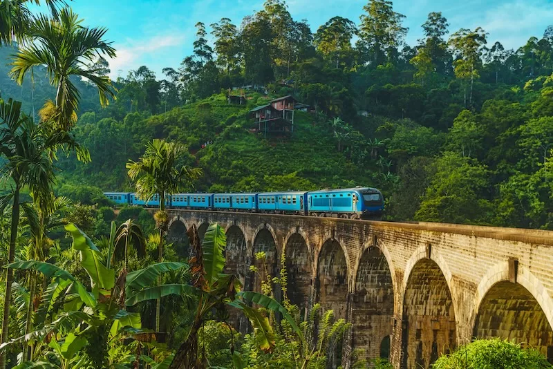 A blue train crosses the iconic Nine Arches Bridge in Ella Sri Lanka surrounded by lush green tea plantations and tropical vegetation on misty forested hills