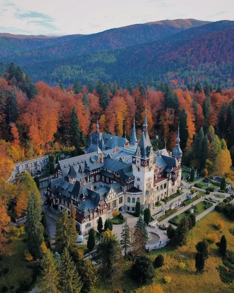An aerial view of the stunning Peles Castle in Sinaia, Romania, nestled among vibrant autumn forests with orange and red trees against a mountain backdrop.