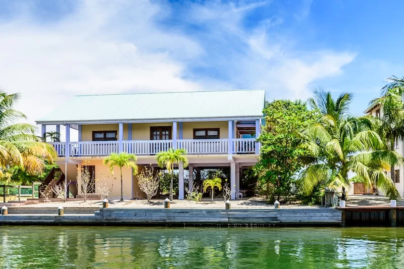 A beautiful two-story tropical home with a wide balcony sits on stilts over a calm canal in Belize, surrounded by lush palm trees and a clear blue sky. 
