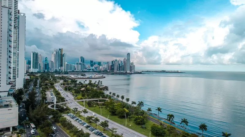 A panoramic daytime view of Panama City's modern skyline along the coast, with a busy highway, green parks, and the vast Pacific Ocean under a partly cloudy blue sky. 
