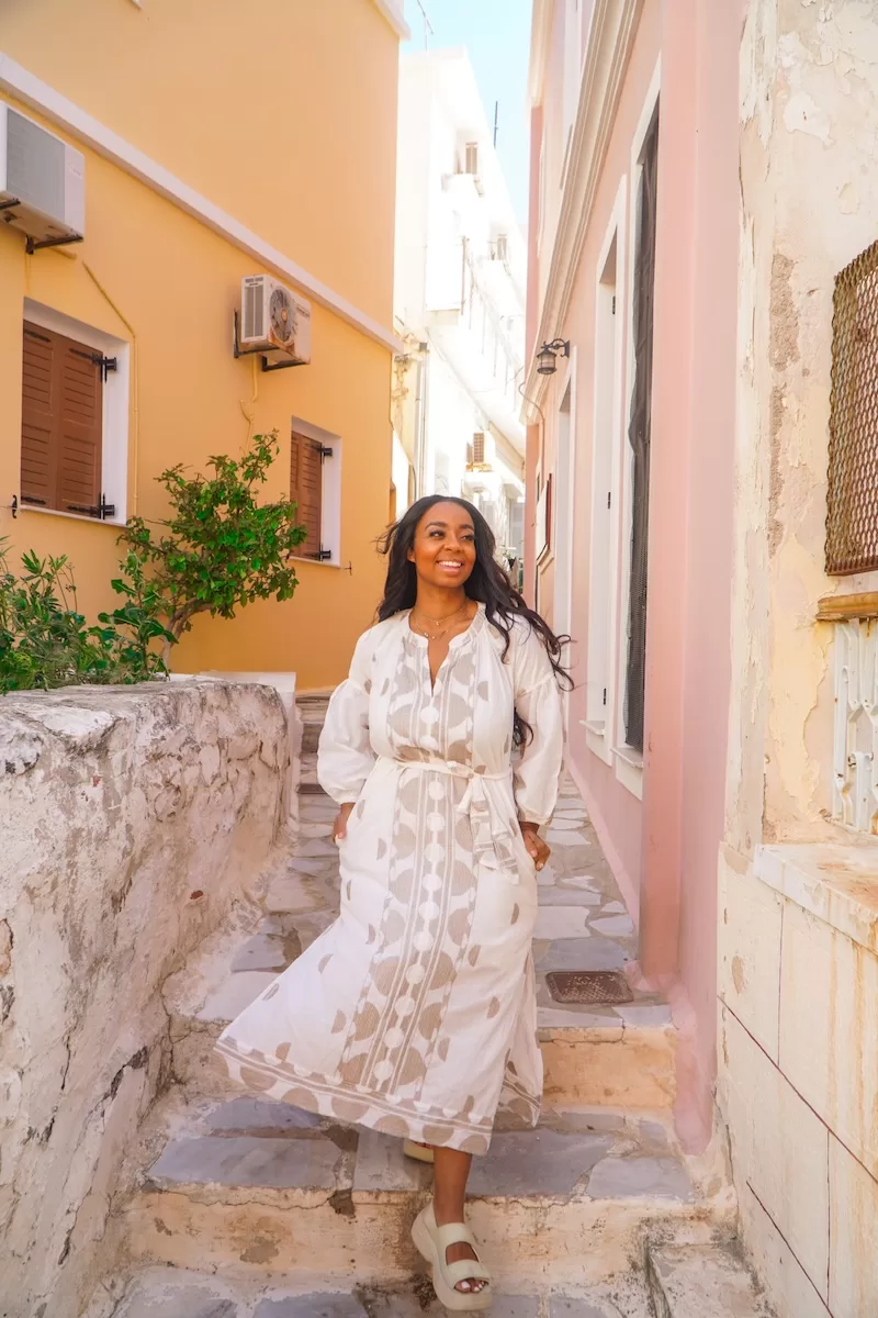 A woman in a flowing white patterned dress smiles warmly while walking down a narrow, sun-drenched alleyway lined with colorful Mediterranean buildings